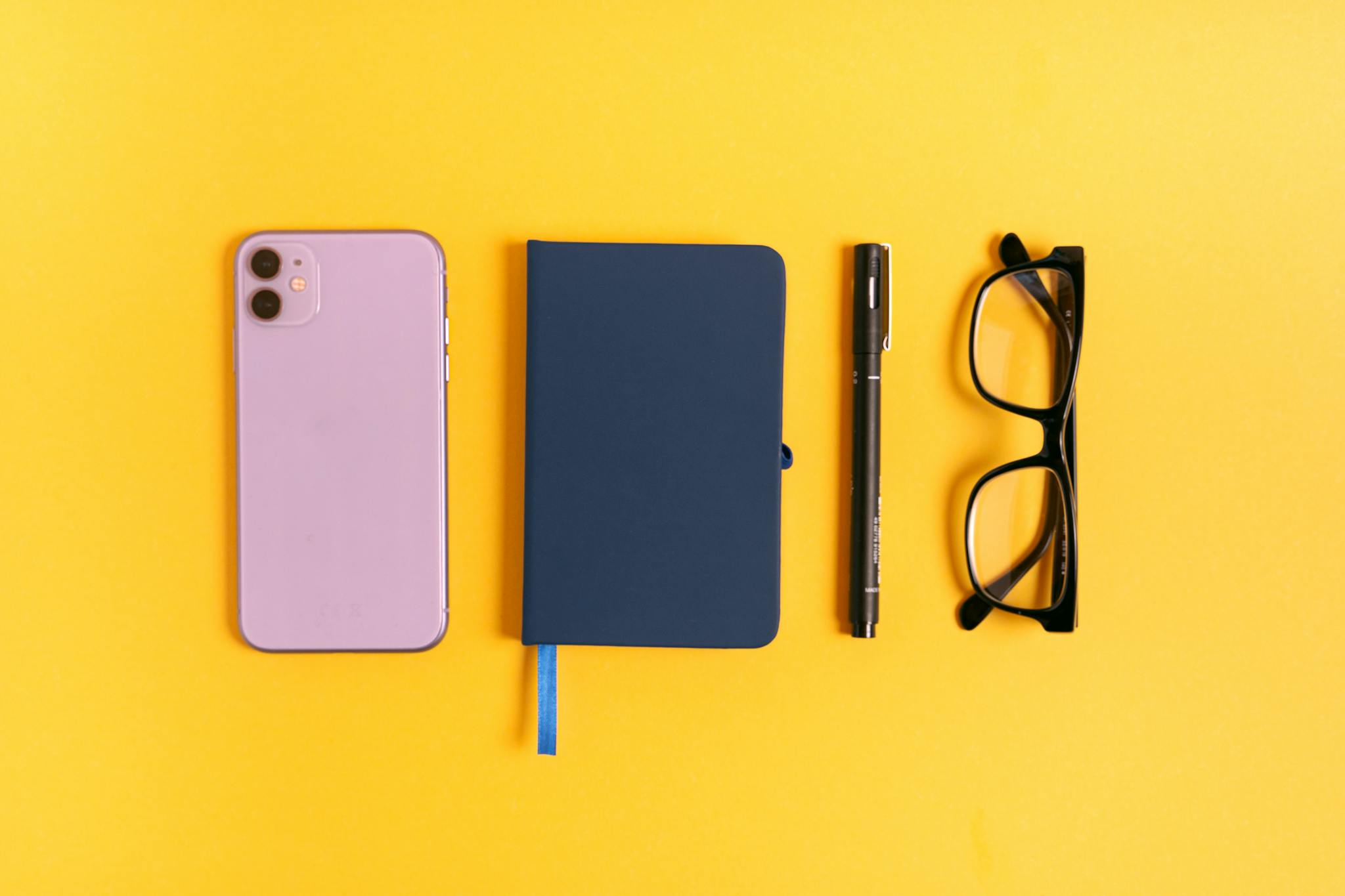 A minimalist flat lay featuring a smartphone, notebook, pen, and glasses on a vibrant yellow background.